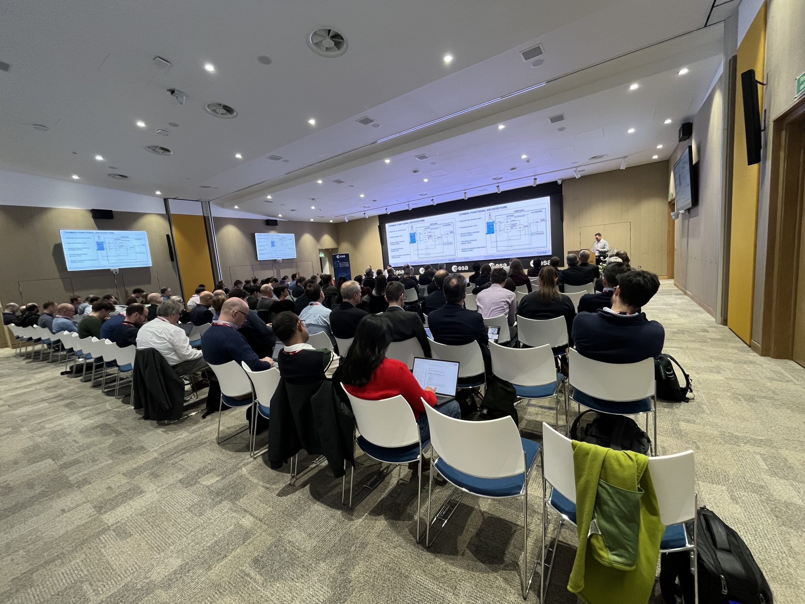 Room of people in a lecture theatre watching a man presenting. There are two large screens